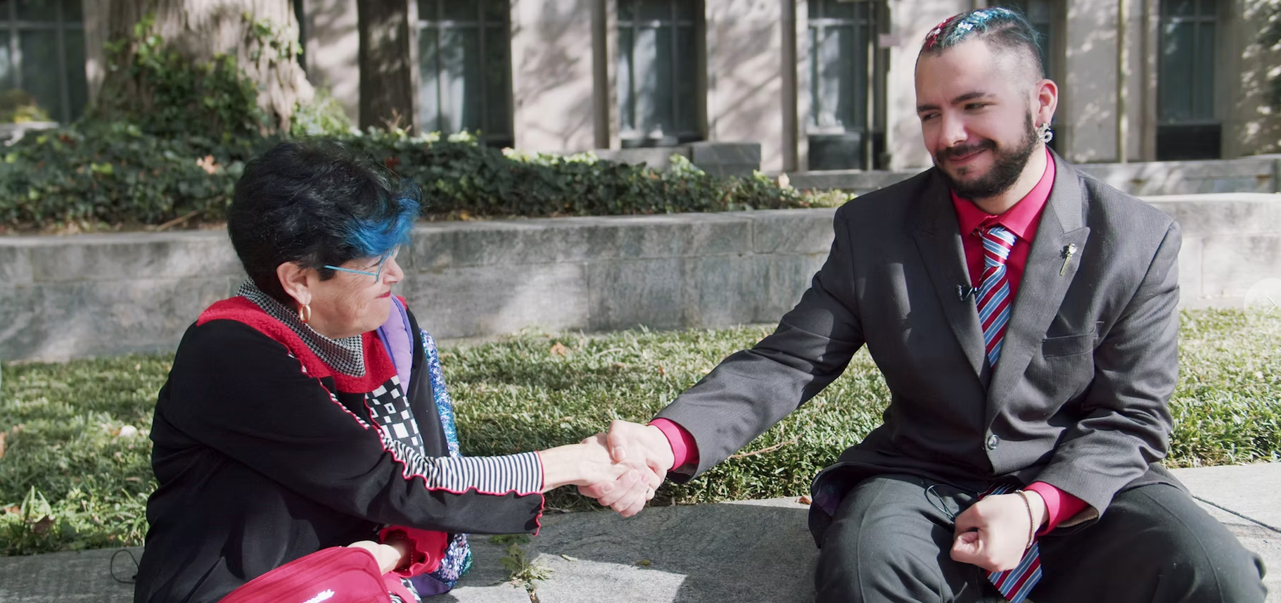 A white woman with black hair and a blue streak of bang wearing a black jacket with red accents shakes hands with a white man with brown hair, bread and mustache wearing a gray suit